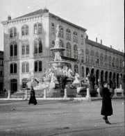 Ein großer Platz mit einem monumentalen Brunnen im Zentrum, umgeben von mehrstöckigen Gebäuden mit Arkadengängen im österreichisch-ungarischen Baustil. Zwei Personen in dunkler Kleidung sind auf dem gepflasterten Platz zu sehen.