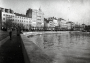 Die Aufnahme zeigt eine mediterrane Hafenpromenade mit einer Reihe mehrstöckiger Gebäude entlang des Wassers. Auf dem Kai sind Personen zu sehen, und die Architektur deutet auf einen österreichisch-ungarischen oder italienischen Hafen hin.