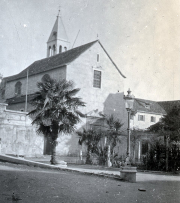 Eine Kirche mit charakteristischem Glockenturm und Satteldach steht zwischen Palmen und südländischer Vegetation. Die mediterrane Architektur und die Palmen deuten auf eine Küstenstadt in der Adria-Region hin.
