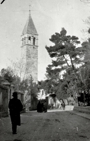Die Aufnahme zeigt einen markanten Kirchturm mit spitzem Dach und Glockengeschoss in einer südeuropäischen Stadt. Auf der Straße sind mehrere Personen zu sehen, darunter eine uniformierte Gestalt im Vordergrund.