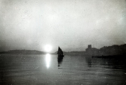 Ein Segelboot mit dunklem Segel fährt bei Sonnenuntergang durch eine ruhige Bucht. Im Hintergrund sind Küstenformationen und Gebäude zu erkennen, während sich das Sonnenlicht auf der Wasseroberfläche spiegelt.