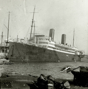 Ein großer Passagierdampfer mit zwei charakteristischen Schornsteinen liegt im Hafen vor Anker. Im Vordergrund sind Hafenausrüstung und Taue zu sehen, während weitere Schiffe mit Masten im Hintergrund erkennbar sind.