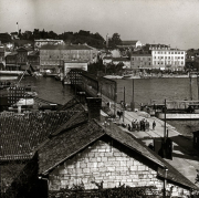 Blick über eine mediterrane Hafenstadt mit mehrstöckigen Gebäuden und bewaldeten Hügeln im Hintergrund. Im Hafen sind mehrere Kriegsschiffe und Marineeinrichtungen zu erkennen, während Marinepersonal auf den Kais versammelt ist.