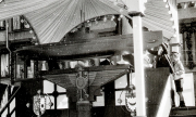 In einem festlich mit Girlanden und Wappen geschmückten Saal steht ein großes Schiffsmodell auf einem Podest. Mehrere Marineoffiziere in Uniform sind bei der zeremoniellen Veranstaltung anwesend.