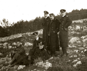Sechs Offiziere der Kaiserlichen Marine und österreichische Militärs posieren in steiniger Küstenlandschaft während eines Landausflugs. Die Aufnahme zeigt Uniformträger verschiedener Ränge in entspannter Haltung zwischen Felsen und niedrigem Bewuchs.