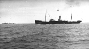 Ein großes Dampfschiff mit charakteristischem Schornstein und Masten fährt auf ruhiger See. Im Hintergrund ist ein weiteres Schiff zu erkennen, während am Himmel ein Flugzeug zu sehen ist.