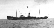 Ein großes Dampfschiff mit charakteristischem Schornstein und zwei Masten fährt auf ruhiger See. Über dem Schiff ist ein Flugzeug am Himmel zu erkennen.