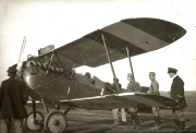Ein deutsches Doppeldecker-Militärflugzeug steht auf einem Flugplatz, während mehrere Soldaten und Offiziere in Uniformen um das Flugzeug versammelt sind. Das Flugzeug zeigt die typische Bauweise der Ersten Weltkriegszeit mit Doppeldeckerflügeln und Propellerantrieb.