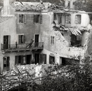 Ein mehrstöckiges Wohngebäude zeigt massive Kriegsschäden mit eingestürzten Dachteilen und zerstörten Fassadenbereichen. Die Fensterläden und der eiserne Balkon sind teilweise erhalten geblieben, während Trümmer die Umgebung bedecken.