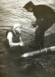 Ein Matrose der Kaiserlichen Marine schwimmt mit einem Tauchretter im Wasser neben dem Deck eines U-Bootes. Ein Besatzungsmitglied auf dem Deck beobachtet die Übung oder Rettungsmaßnahme.