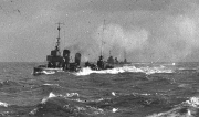 Ein deutsches Kriegsschiff mit mehreren Schornsteinen und Aufbauten fährt bei bewegter See mit hoher Geschwindigkeit. Das Schiff erzeugt eine deutlich sichtbare Bugwelle und Gischt.