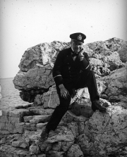Ein Offizier der Kaiserlichen Marine in dunkler Uniform mit Schirmmütze sitzt auf einem Felsvorsprung an einer zerklüfteten Küste. Die mediterrane Landschaft und das Meer im Hintergrund deuten auf den Operationsraum der deutschen U-Boot-Flottille im Mittelmeer hin.