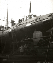 Ein deutsches U-Boot liegt im Trockendock einer Werft, umgeben von Gerüsten und Arbeitsplattformen. Besatzungsmitglieder und Werftarbeiter sind auf dem Boot und in der Dockanlage bei Wartungs- oder Reparaturarbeiten zu sehen.
