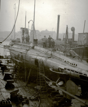 Ein deutsches U-Boot liegt im Trockendock einer Werft, umgeben von Kränen und Werftanlagen. Zwei Besatzungsmitglieder in Uniformen stehen auf dem Kommandoturm des Bootes.