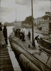 Ein deutsches U-Boot liegt vertäut an einem Hafenpier, während Besatzungsmitglieder auf dem Deck arbeiten. Im Hintergrund sind Hafengebäude und weitere Marineeinrichtungen zu erkennen.