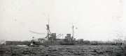 Ein großes Schlachtschiff der deutschen Kaiserlichen Marine liegt im Hafen, erkennbar an den charakteristischen Geschütztürmen und der militärischen Bauweise. Im Vordergrund ist ein kleineres Fahrzeug auf dem Wasser zu sehen.