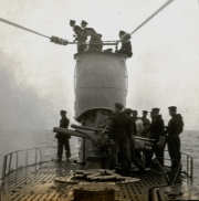 Marineangehörige in Uniform versammeln sich um ein Deckgeschütz auf einem deutschen Kriegsschiff. Die Aufnahme zeigt den Schornstein und die Takelage des Schiffes bei ruhiger See.
