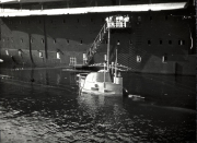Ein U-Boot liegt in einem überdachten Hafendock, während mehrere Besatzungsmitglieder auf einer Gangway über dem Turm stehen. Die Aufnahme zeigt die typische Infrastruktur einer U-Boot-Basis mit massiven Betonwänden und Fenstern.