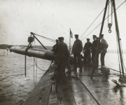 Die Besatzung des deutschen U-Bootes S.M. U 39 übernimmt mit Hilfe eines Krans einen Torpedo auf hoher See. Mehrere Marineoffiziere und Matrosen in Uniformen stehen auf dem nassen Deck und überwachen den Ladevorgang.