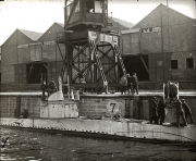 Ein deutsches U-Boot liegt in einem Trockendock einer Werft, umgeben von Backsteingebäuden und einem Kran. Mehrere Besatzungsmitglieder und Werftarbeiter stehen auf dem Deck und am Kai.
