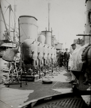 Mehrere Seeminen sind auf dem Deck eines Kriegsschiffes aufgereiht, während Marineoffiziere in Uniformen daneben stehen. Die zylindrischen Minen mit ihren charakteristischen Kontakthörnern sind zur Verladung oder Installation bereit.