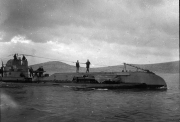 Ein deutsches U-Boot der Kaiserlichen Marine schwimmt aufgetaucht in ruhiger See, während mehrere Besatzungsmitglieder auf dem Deck und am Turm stehen. Im Hintergrund ist eine hügelige Küstenlandschaft unter bewölktem Himmel zu erkennen.