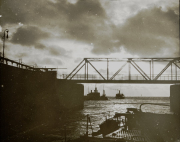 Mehrere Kriegsschiffe der Kaiserlichen Marine liegen unter einer großen Eisenbahnbrücke in einem Hafen vor Anker. Die Aufnahme zeigt die charakteristische Silhouette von Torpedobooten oder kleineren Kriegsschiffen bei bewölktem Himmel.