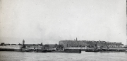 Die Aufnahme zeigt eine befestigte Hafenanlage mit Molen, Kränen und militärischen Einrichtungen vor einer Festung auf einem Hügel. Im Vordergrund sind Hafenbecken und Pier-Anlagen der Kaiserlichen Marine zu erkennen.