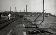 Blick auf eine Hafenanlage mit hölzernen Stegen und Anlegevorrichtungen, auf denen Marinepersonal in weißer Uniform zu sehen ist. Im Hintergrund erstreckt sich eine Küstenstadt mit Gebäuden und Industrieanlagen auf den Hügeln.