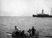 Ein Rettungsboot mit mehreren Personen schwimmt auf ruhiger See, während im Hintergrund ein großes Dampfschiff mit Schornstein sichtbar ist. Die Aufnahme dokumentiert eine typische Szene des U-Boot-Krieges, bei der Überlebende eines versenkten Schiffes gerettet werden.