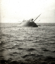 Ein Dampfschiff sinkt mit stark geneigter Backbordlage und ragenden Masten aus dem bewegten Meer. Die Aufnahme dokumentiert eine Schiffsversenkung durch S.M. U 39 während des U-Boot-Krieges.
