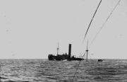Ein Dampfschiff mit charakteristischem Schornstein und zwei Masten liegt schwer beschädigt oder sinkend im Wasser. Die Aufnahme erfolgte von einem anderen Schiff aus, dessen Takelage im Vordergrund sichtbar ist.