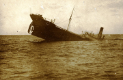 Ein Dampfschiff mit deutlicher Schlagseite nach Backbord sinkt langsam ins Meer. Das Schiff zeigt noch Masten und Schornstein, während es bereits tief im Wasser liegt.