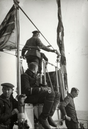 Mehrere Marineoffiziere und Besatzungsmitglieder der Kaiserlichen Marine auf der Brücke eines Kriegsschiffs. Ein Offizier mit Mütze steht am Mast und bedient nautische Ausrüstung, während andere Besatzungsmitglieder in Uniformen verschiedene Positionen auf der Brücke einnehmen.