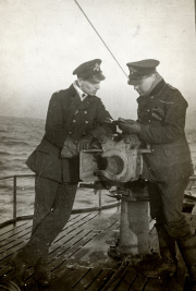 Zwei Offiziere der Kaiserlichen Marine in Uniformen und Schirmmützen bedienen gemeinsam einen Sextanten auf dem Holzdeck eines Kriegsschiffs. Die Aufnahme zeigt die Navigation auf See während des Ersten Weltkriegs.