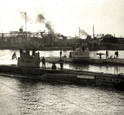 Mehrere deutsche U-Boote liegen im Hafenbecken vor Anker, während Besatzungsmitglieder auf den Decks zu sehen sind. Im Hintergrund sind Werftanlagen mit Kränen und Industriebauten sowie aufsteigender Rauch erkennbar.