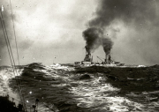 Ein deutsches Schlachtschiff der Kaiserlichen Marine fährt bei schwerem Seegang und stößt dunkle Rauchsäulen aus seinen Schornsteinen aus. Die Aufnahme wurde von einem anderen Schiff aus gemacht, dessen Takelage am linken Bildrand sichtbar ist.