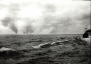 Ein U-Boot beobachtet die Versenkung eines Schiffes, aus dem mehrere dunkle Rauchsäulen aufsteigen. Die Aufnahme zeigt die typische Kriegsführung deutscher U-Boote im Ersten Weltkrieg gegen Handelsschiffe.