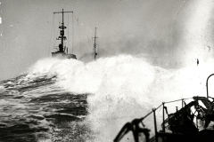 Ein Kriegsschiff kämpft sich durch schwere See, während hohe Wellen über Bug und Aufbauten brechen. Die Aufnahme wurde von einem anderen Schiff aus gemacht, dessen Deck und Ausrüstung im Vordergrund zu sehen sind.