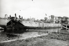 Ein stark beschädigtes Kriegsschiff liegt im Hafen, wobei mehrere Besatzungsmitglieder auf dem Deck stehen. Das Schiff weist schwere Zerstörungen am Heck und an den Aufbauten auf.