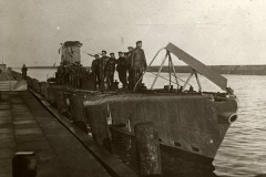 Ein deutsches U-Boot liegt im Hafen, während die Besatzung in Uniformen auf dem Deck und Turm versammelt ist. Das Foto zeigt das Fahrzeug von der Seite mit deutlich sichtbarem Kommandoturm und Periskop.