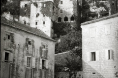 Die Aufnahme zeigt eine mediterrane Küstenstadt mit charakteristischer Steinarchitektur, einem markanten Kirchturm und Pferdewagen im Vordergrund. Die terrassenförmig an einem Hang gebauten Gebäude und der architektonische Stil deuten auf die Adria-Region hin.