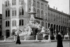 Ein großer Platz mit einem monumentalen Brunnen im Zentrum, umgeben von mehrstöckigen Gebäuden mit Arkadengängen im österreichisch-ungarischen Baustil. Zwei Personen in dunkler Kleidung sind auf dem gepflasterten Platz zu sehen.