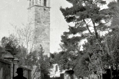 Die Aufnahme zeigt einen markanten Kirchturm mit spitzem Dach und Glockengeschoss in einer südeuropäischen Stadt. Auf der Straße sind mehrere Personen zu sehen, darunter eine uniformierte Gestalt im Vordergrund.