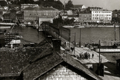 Blick über eine mediterrane Hafenstadt mit mehrstöckigen Gebäuden und bewaldeten Hügeln im Hintergrund. Im Hafen sind mehrere Kriegsschiffe und Marineeinrichtungen zu erkennen, während Marinepersonal auf den Kais versammelt ist.