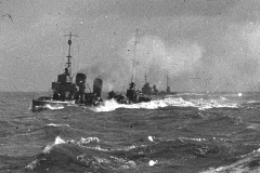 Ein deutsches Kriegsschiff mit mehreren Schornsteinen und Aufbauten fährt bei bewegter See mit hoher Geschwindigkeit. Das Schiff erzeugt eine deutlich sichtbare Bugwelle und Gischt.