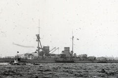 Ein großes Schlachtschiff der deutschen Kaiserlichen Marine liegt im Hafen, erkennbar an den charakteristischen Geschütztürmen und der militärischen Bauweise. Im Vordergrund ist ein kleineres Fahrzeug auf dem Wasser zu sehen.