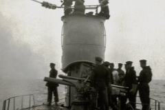 Marineangehörige in Uniform versammeln sich um ein Deckgeschütz auf einem deutschen Kriegsschiff. Die Aufnahme zeigt den Schornstein und die Takelage des Schiffes bei ruhiger See.