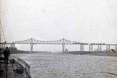 Die Aufnahme zeigt eine monumentale Eisenbahnbrücke mit charakteristischer Stahlkonstruktion, fotografiert vom Deck eines Kriegsschiffes aus. Ein Matrose in dunkler Uniform steht am linken Bildrand auf dem Schiffsdeck.