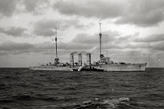 Ein deutscher Kreuzer mit drei charakteristischen Schornsteinen fährt bei bewegter See unter bewölktem Himmel. Das Kriegsschiff zeigt die typische Silhouette eines Kreuzers der Kaiserlichen Marine aus der Zeit des Ersten Weltkriegs.